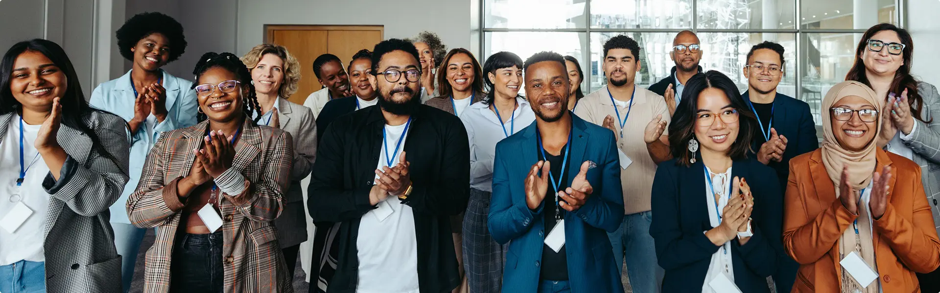 A diverse group of people wearing conference badges stand together indoors, smiling and applauding toward the camera.